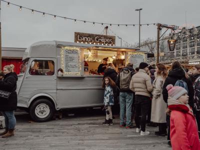 People gather at a cozy Christmas market in Zürich, enjoying crepes from a vintage food truck adorned with lights.