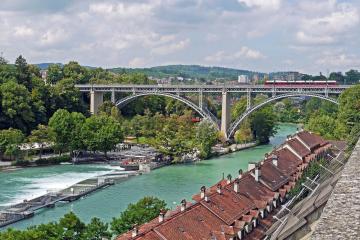 Panoramic view of the historic Kirchenfeld Bridge over the Aare River in Bern, surrounded by lush greenery and traditional rooftops.
